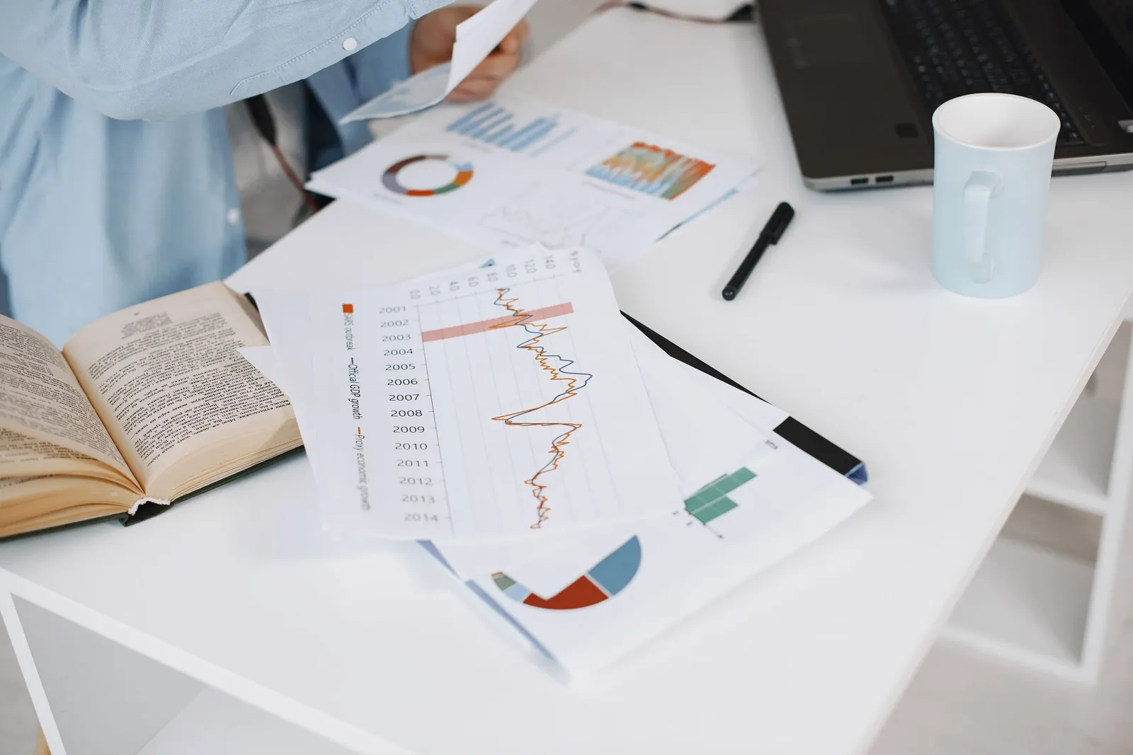 Printed charts and an open book lie on a white desk beside a laptop and mug.