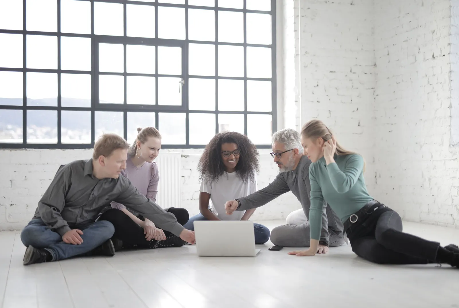Five adults of different ages sit on the floor of a bright room, working together around a laptop.