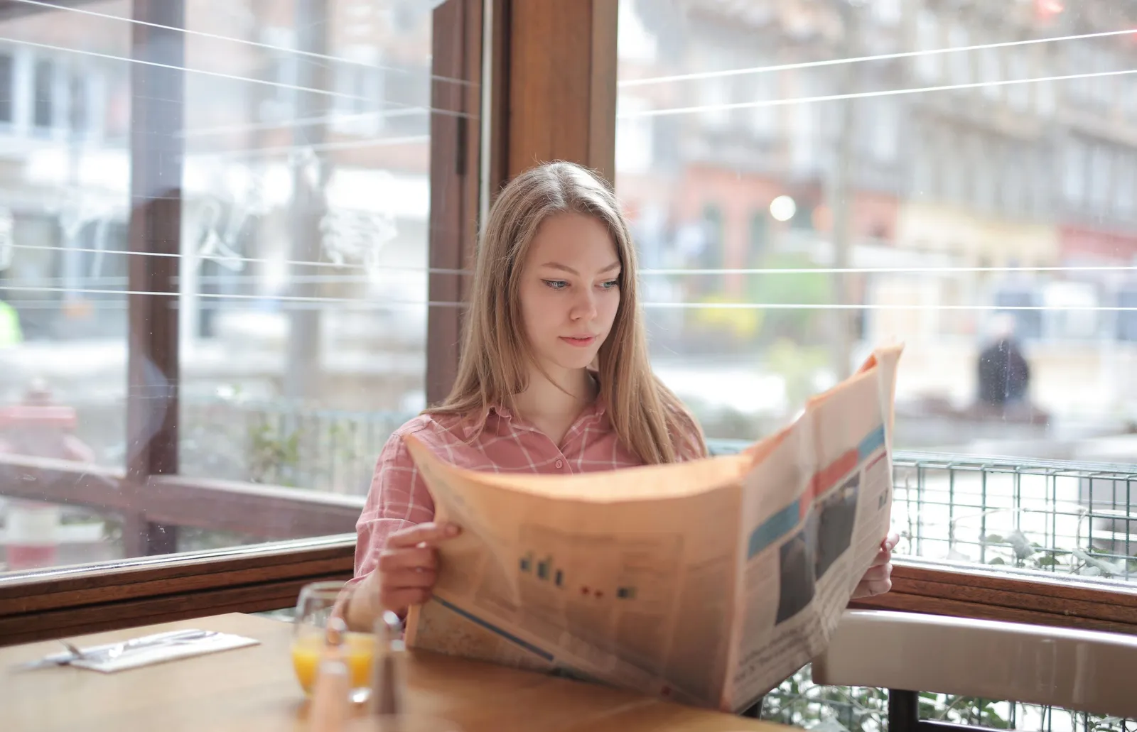 A young woman reads a newspaper at a sunny cafe window.