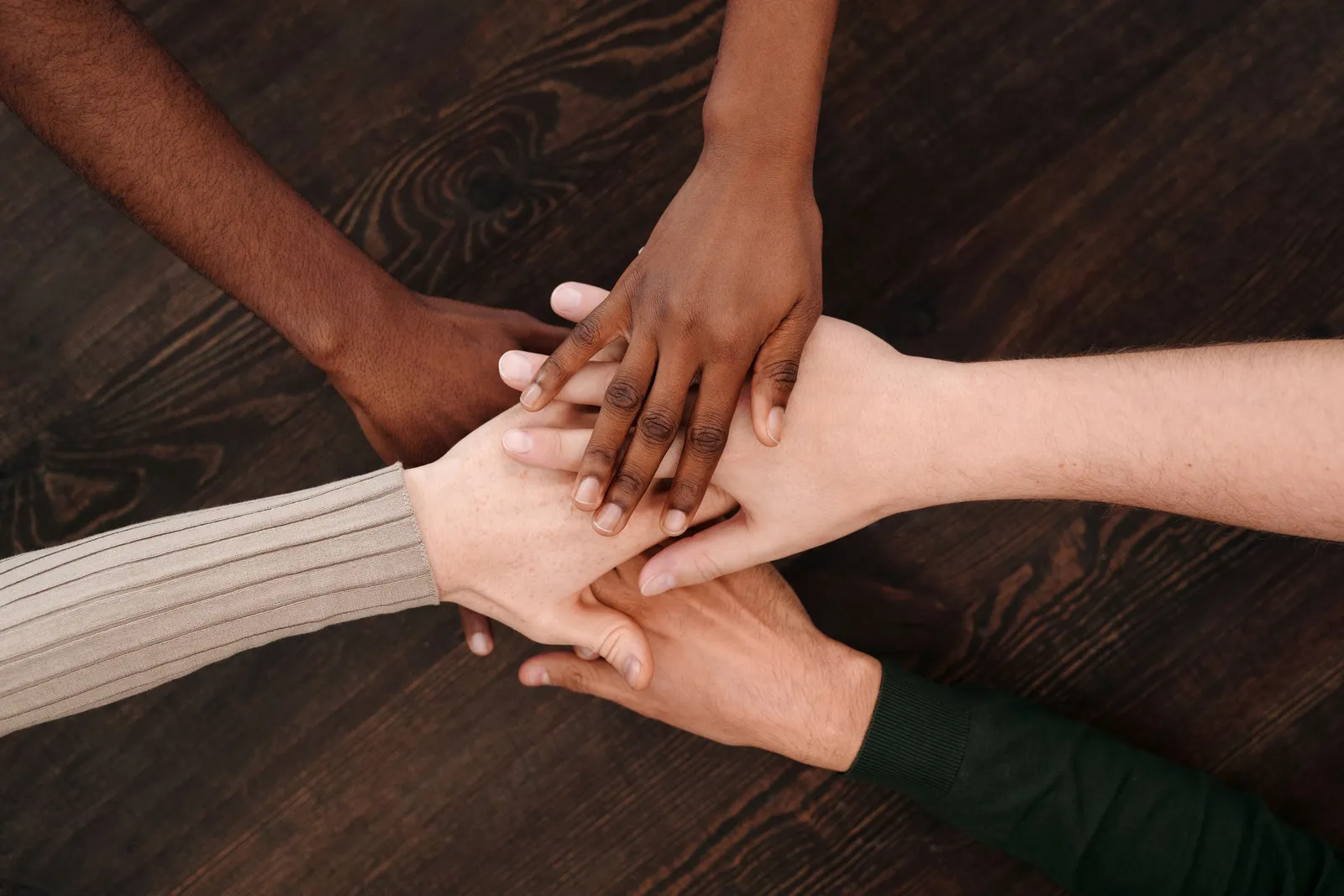 Hands of people with different skin tones rest on top of one another on a wooden table.