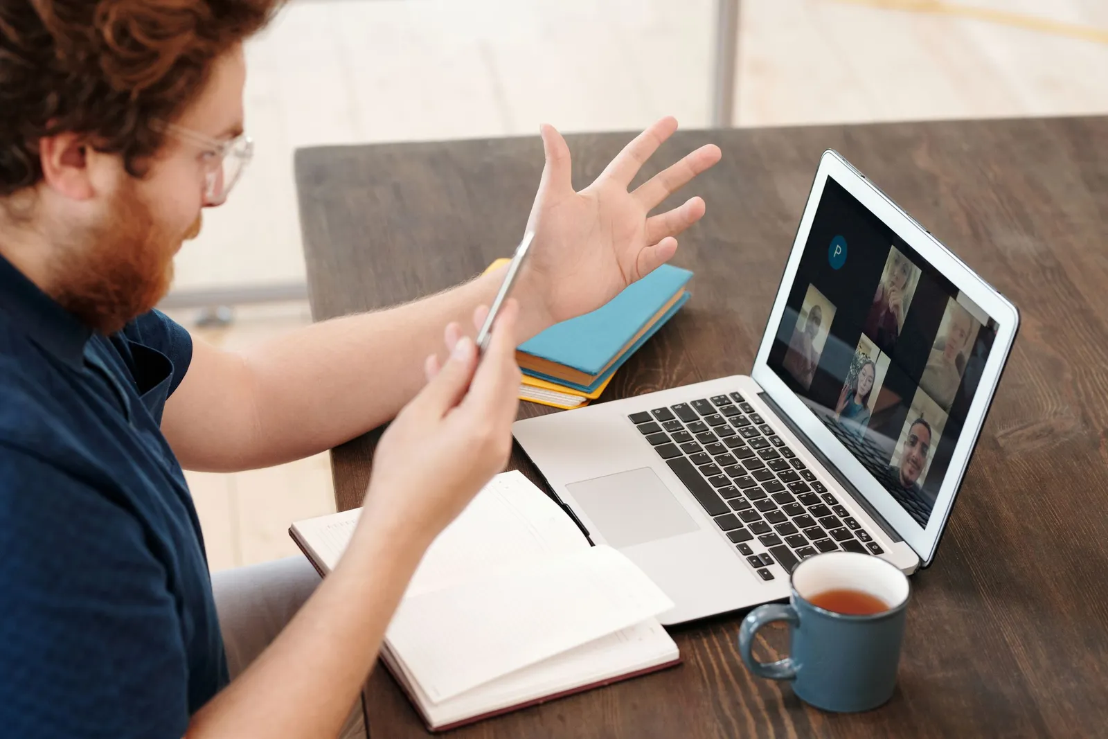 A man with a mug of tea and an open notebook gestures while chatting with a gallery of participants on a laptop video call.