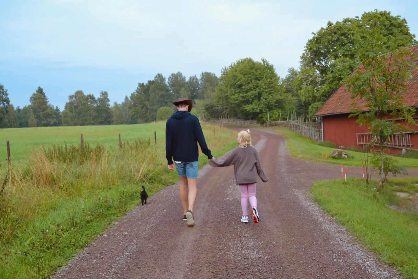 Seen from behind, an adult and a young child walk hand in hand down a country road past a red barn.