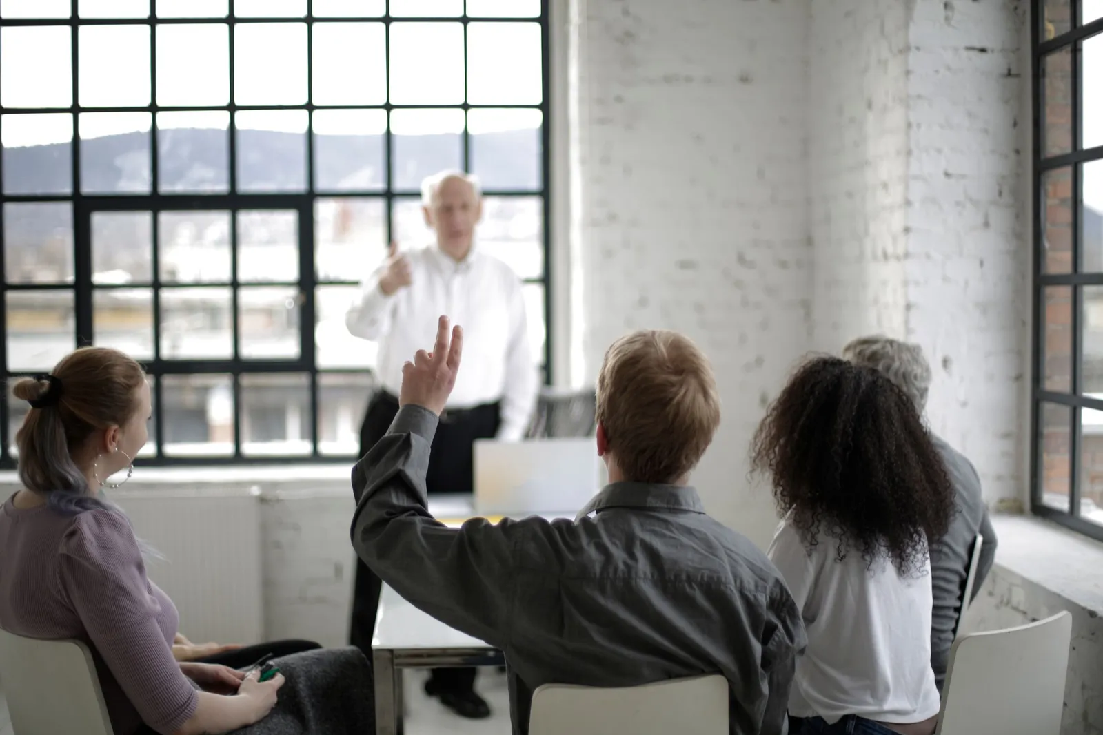 Seen from behind, a participant raises a hand while an instructor leads a session in a bright room.