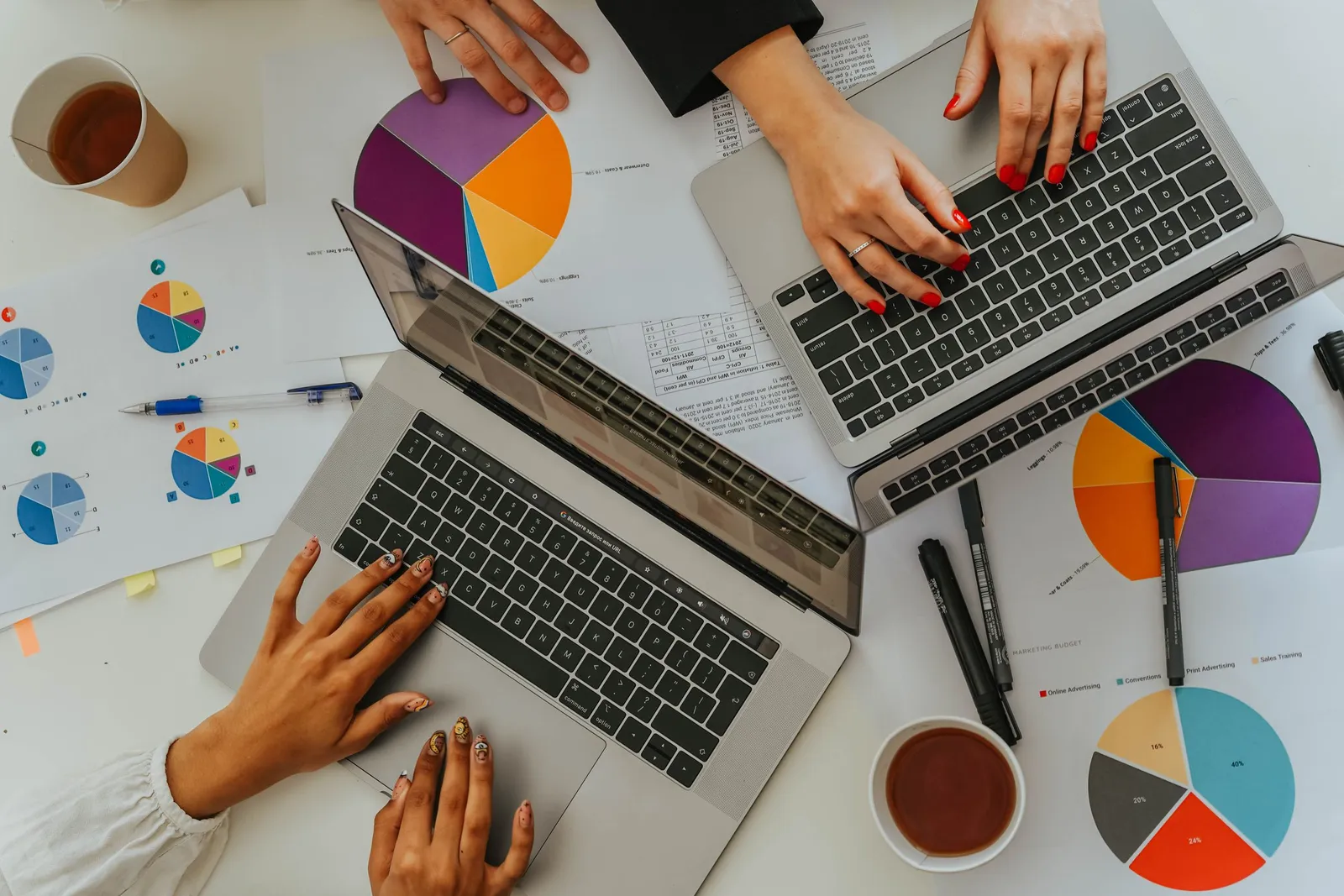 Seen from above, two people work on laptops among printed pie charts and cups of tea.