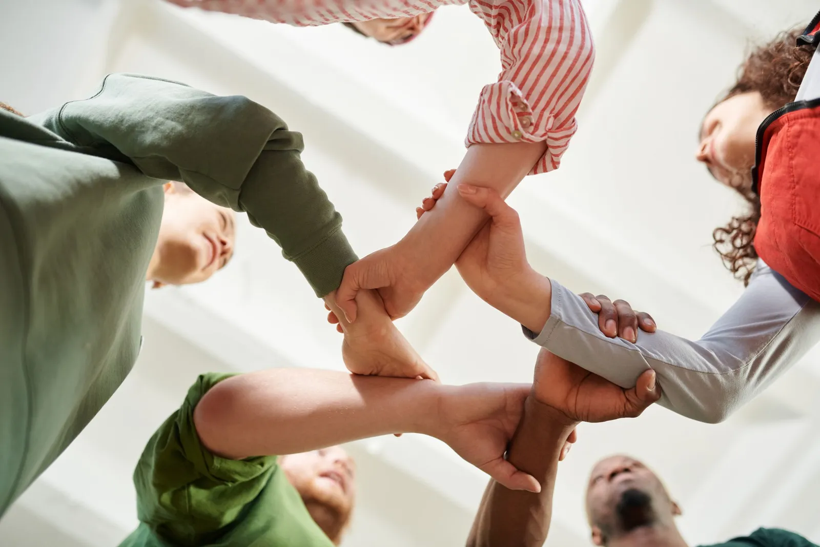 Seen from below, five people join hands in a circle against a bright ceiling.